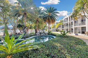 A resort with a swimming pool surrounded by tropical plants and palm trees, with a building featuring balconies in the background.