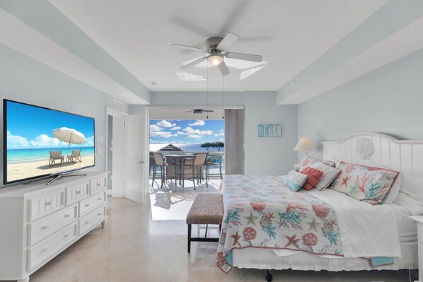 A bedroom with a bed, a TV, a ceiling fan, and a view of the beach.