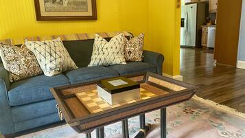 A living room with a blue sofa, patterned cushions, a chessboard on a woven coffee table, and a framed picture on the wall.