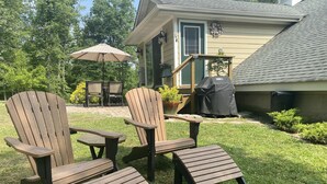 Un espacio al aire libre con sillas Adirondack y una mesa, una parrilla cubierta y una casa con porche.
