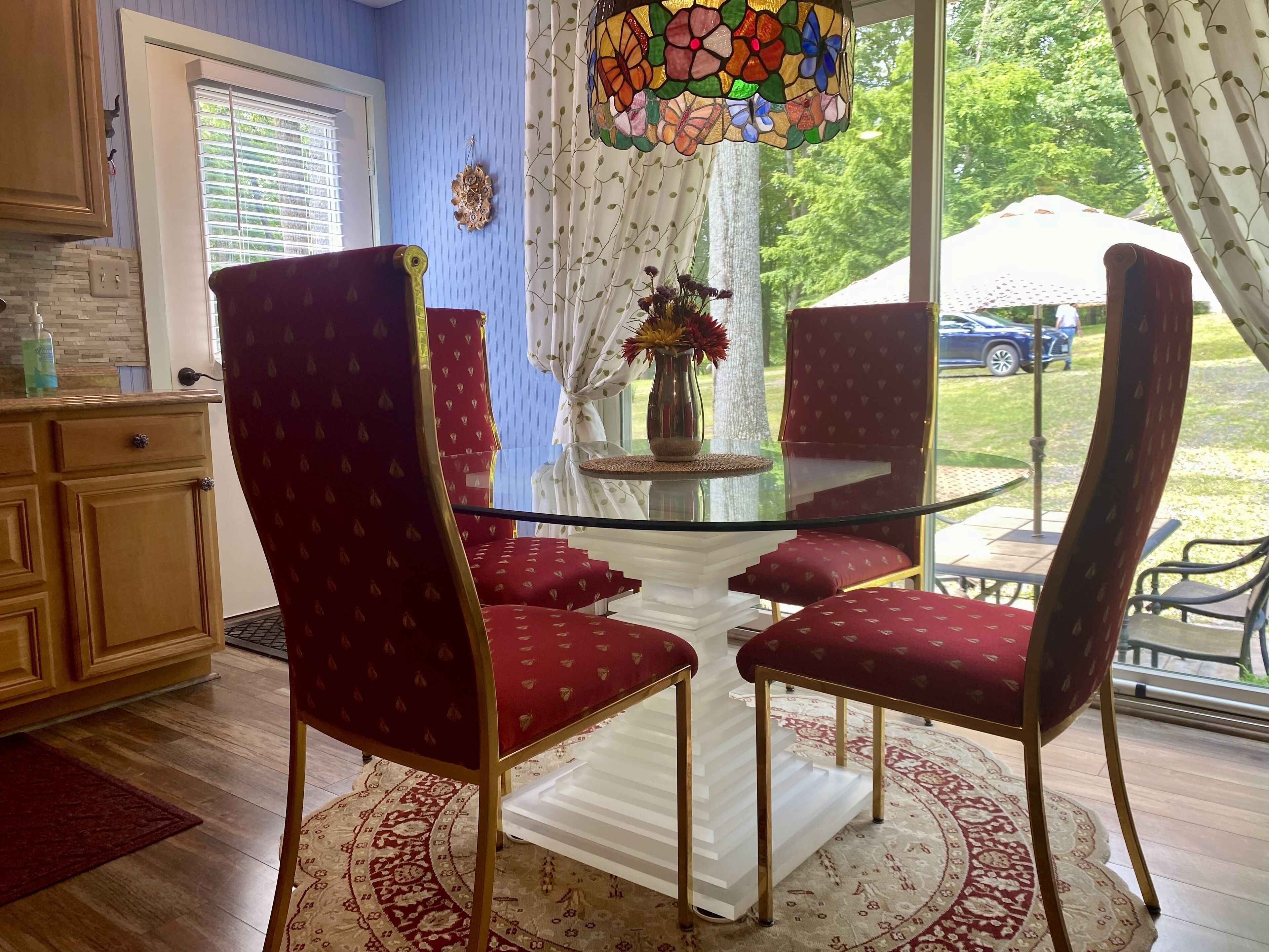 A dining area with a glass table surrounded by red upholstered chairs, a decorative vase with flowers, and a stained glass window above.