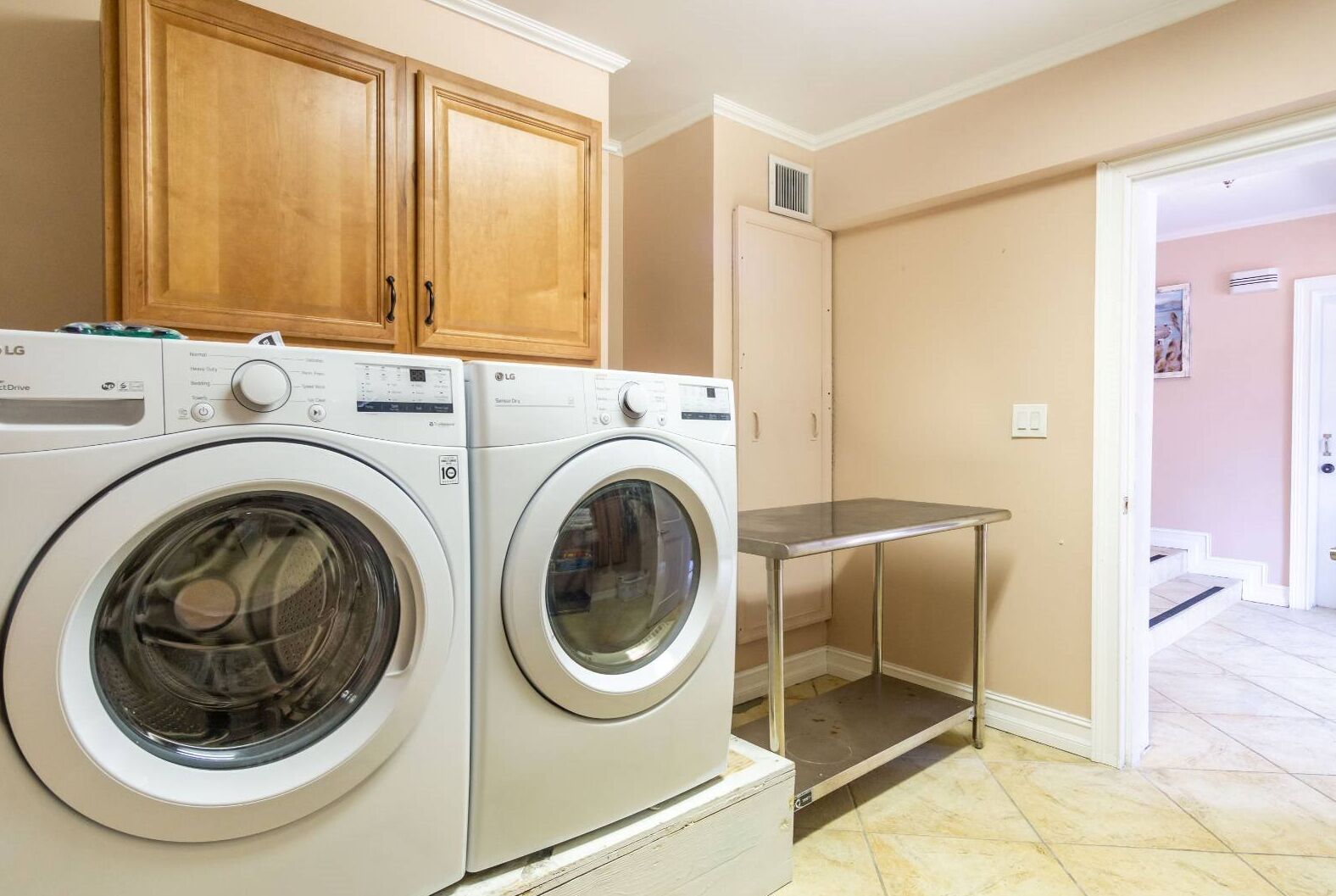A laundry area with two washing machines, wooden cabinets, a metal table, and a door leading to another room.