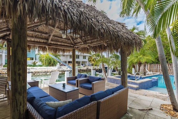 A poolside area with wicker furniture, a thatched roof, and a view of a pool and resort buildings.