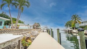 A wooden dock leading to a marina with boats, palm trees, and a thatched-roof structure. - Yellowtail for Dinner – Ground-Level Waterfront 3BR Home with Pool & Dock on the Gulfside (Marathon)