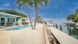 A pool area with palm trees, a dock, and a building with outdoor seating. - Yellowtail for Dinner – Ground-Level Waterfront 3BR Home with Pool & Dock on the Gulfside (Marathon)