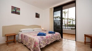 Een slaapkamer met een bed, een nachtkastje en een balkon met uitzicht op palmbomen en gebouwen.