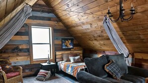 A cozy attic bedroom with a sloped ceiling, a bed with a colorful blanket, a dark armchair, a wicker chair, and a window with curtains. - Balsam Hollow Cabin in the Adirondacks (Bloomingdale)