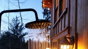 A wooden cabin with a showerhead mounted on a wooden structure, a window with a view of trees, and a warm light fixture on the wall. - Emerald Forest Treehouse - From Treehouse Masters (Redmond)