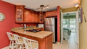 A kitchen with wooden cabinets, a stainless steel refrigerator, a microwave, and a granite countertop. There are three white wicker chairs at the island. - Comfy Condo 1bed/1bath nightly condo with pool (Marathon)