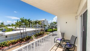 A balcony with two chairs and a small table, overlooking a parking area and a building with palm trees. - Comfy Condo 1bed/1bath nightly condo with pool (Marathon)