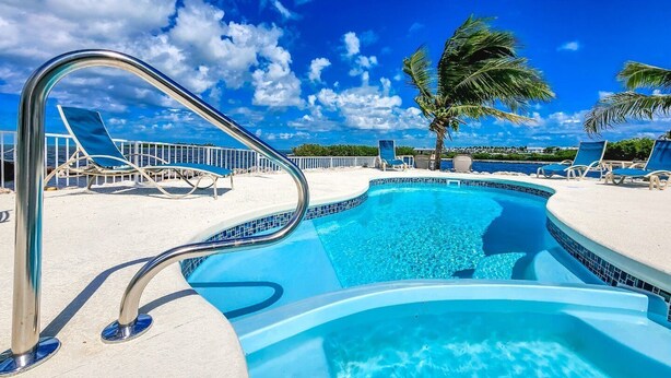 A swimming pool with a metal ladder, surrounded by lounge chairs and palm trees, under a clear blue sky. - Palm Point 3bed/2bath single family home with open water views, private pool & dockage (Marathon)