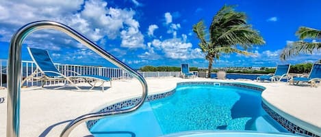 A swimming pool with a metal ladder, surrounded by lounge chairs and palm trees, under a clear blue sky.