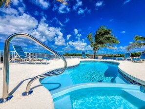 Piscina con scaletta in metallo, circondata da sdraio e palme, sotto un cielo azzurro terso.