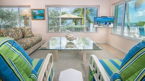 A living room with a glass coffee table, striped armchairs, a sofa, and a view of a tropical landscape through large windows. - Palm Point 3bed/2bath single family home with open water views, private pool & dockage (Marathon)