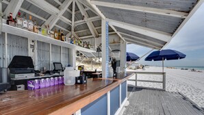 Un bar en la playa con barra de madera, estantes con botellas y sombrillas.