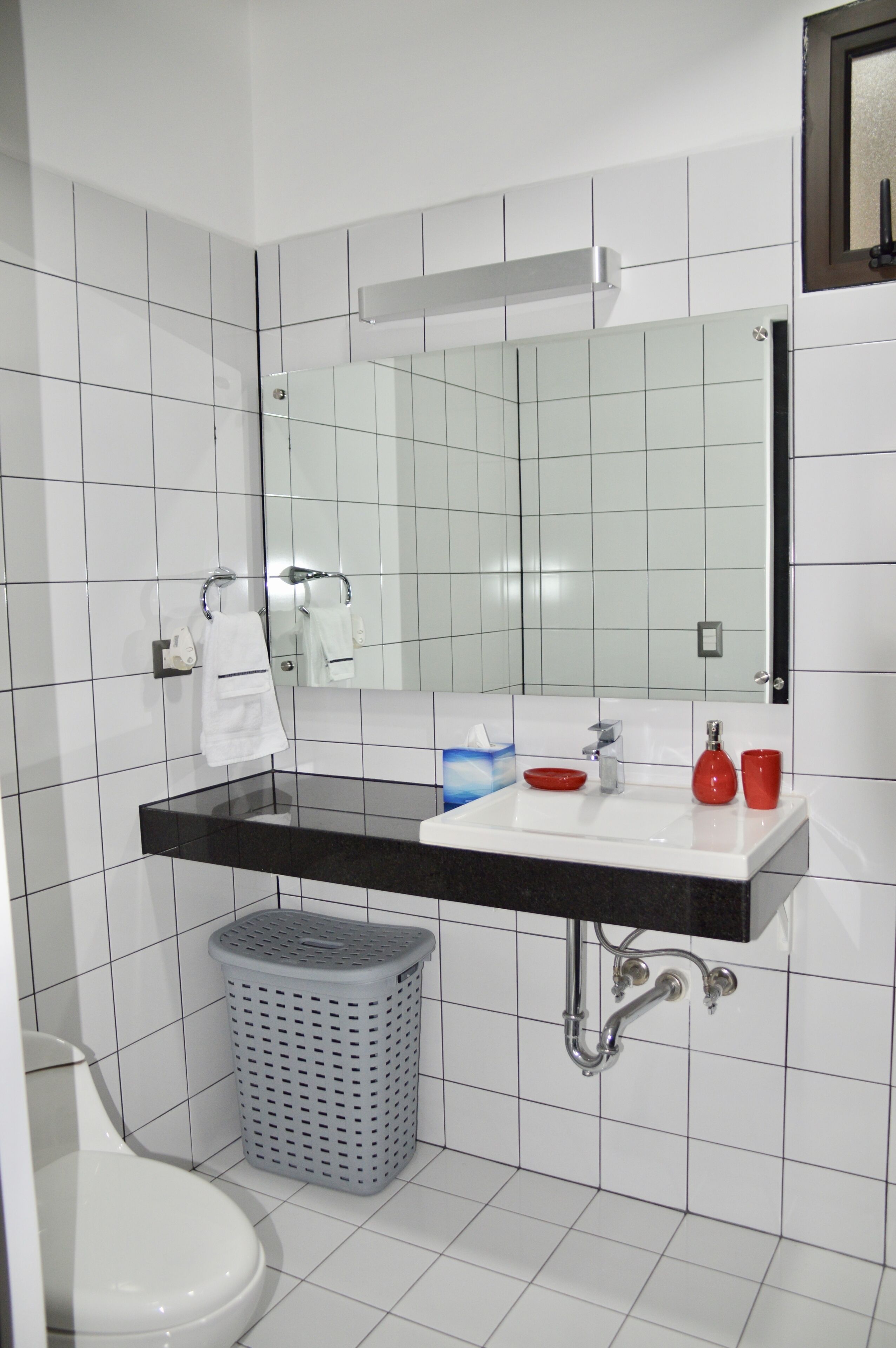 Une salle de bain avec un mur recouvert de carreaux blancs, un plan de toilette noir, un miroir, un lavabo, des toilettes et un panier à linge.