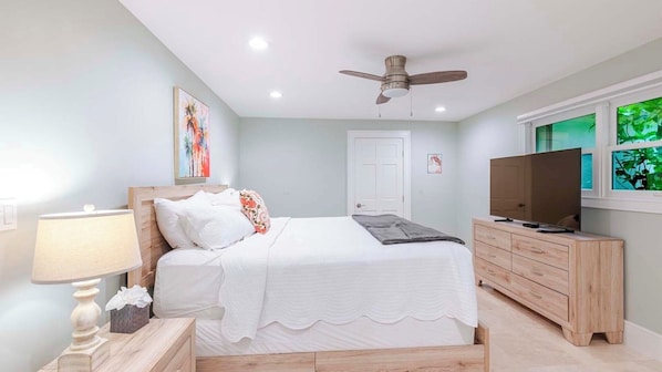 A bedroom with a bed, a ceiling fan, a TV, a dresser, and a window with a view.