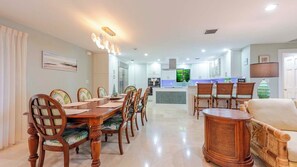 A spacious dining area with a long wooden table and chairs, a kitchen in the background, and a living area with a sofa and a lamp.