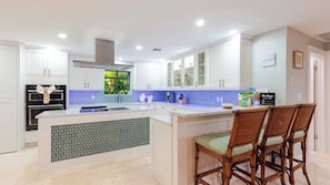 A modern kitchen with white cabinets, a central island, and a dining area with wicker chairs. - Shangri-La 3 Bedroom 3 Baths Gulf Front Estate (Marathon)