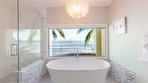 Ein modernes Badezimmer mit einer großzügigen Badewanne, einer Duschkabine aus Glas und Meerblick durch ein großes Fenster.