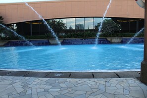 A swimming pool with water jets, surrounded by a stone-tiled walkway and a building with large windows. - Times Square Service Suite By DaisyHomes (Kuala Lumpur)