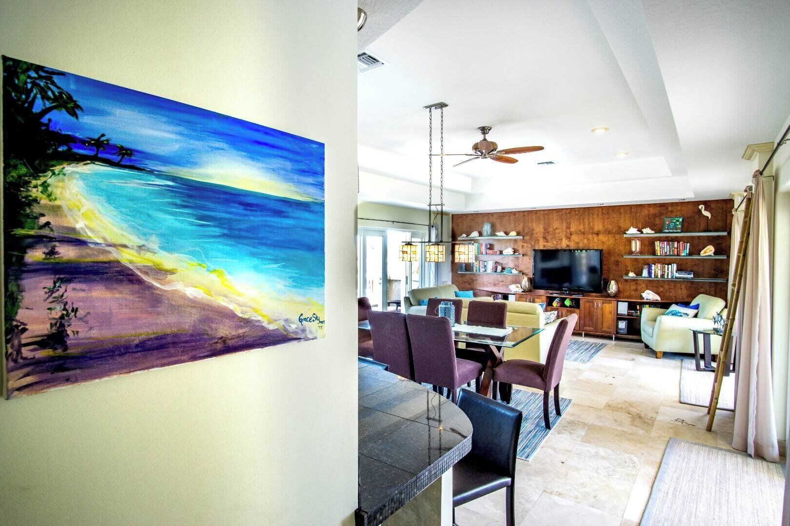 A modern living room with a dining area, a ceiling fan, and a large painting of a beach scene.