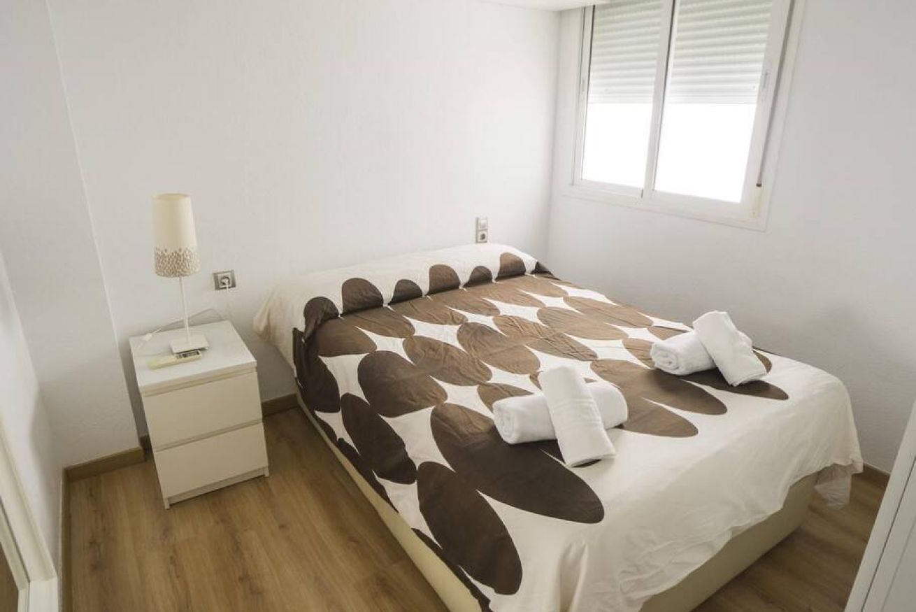 Ein ordentlich bezogenes Bett mit einem gemusterten Überwurf, ein Nachttisch mit einer Lampe und ein Fenster mit Jalousien.