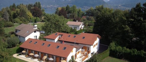 Ein zweistöckiges Gebäude mit rotem Ziegeldach, einer überdachten Außenbereich-Sitzgelegenheit mit Sonnenschirmen und Blick auf Berge im Hintergrund.