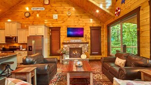 A cozy cabin interior with a fireplace, leather sofas, a TV, wooden walls, and a kitchen area. - Splashin' Around! 3 Bedroom with a Pool! Minutes from downtown Pigeon Forge. (Sevierville)
