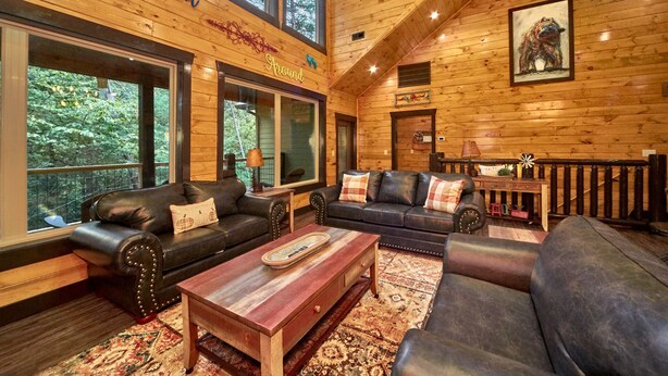 A log cabin interior with two leather sofas, a wooden coffee table, and a large window overlooking greenery. - Splashin' Around! 3 Bedroom with a Pool! Minutes from downtown Pigeon Forge. (Sevierville)