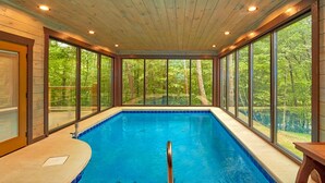 An indoor swimming pool with wooden walls and large windows offering a view of the surrounding forest. - Splashin' Around! 3 Bedroom with a Pool! Minutes from downtown Pigeon Forge. (Sevierville)