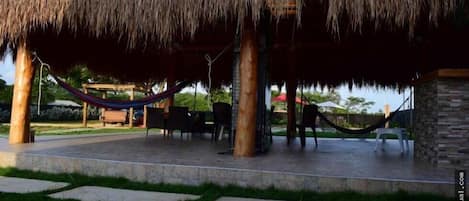 A thatched-roof outdoor seating area with a hammock, stone pillars, and a paved patio.