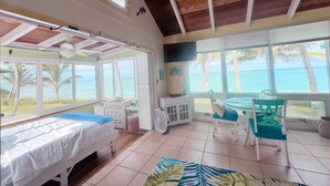 Une chambre avec un lit, un bureau et une chaise, de grandes fenêtres donnant sur la plage et l’océan, et un ventilateur de plafond.