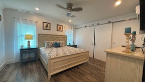 Quarto com cama, mesas de cabeceira, cômoda, ventilador de teto e janela com persianas.