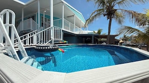 A swimming pool with a white deck, a lifeguard stand, and a house with a blue exterior in the background.