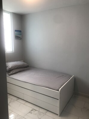 Pieni huone, jossa on yksi sänky, pelkistetty valkoinen seinä, ikkuna, jossa on sälekaihtimet, pieni taulu ja laattalattia.