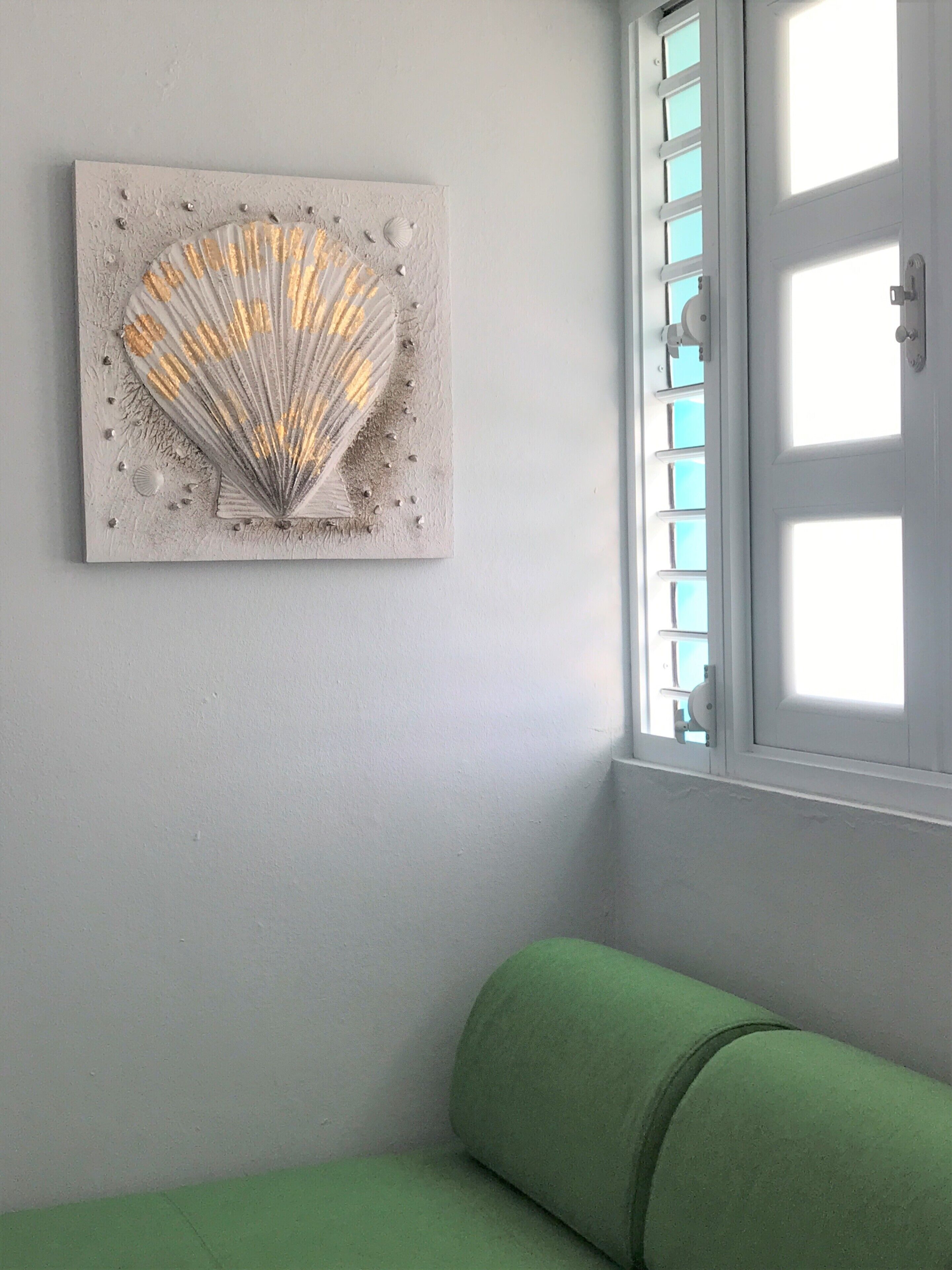 Une pièce avec un canapé vert, une œuvre d’art représentant une coquille accrochée au mur et une fenêtre avec des stores.