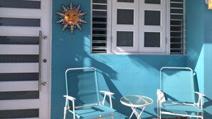 Ein Zimmer mit blauer Wand, einer weißen Tür, einem Fenster mit Rollläden und zwei weißen Stühlen an einem kleinen runden Tisch.