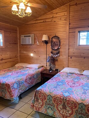 A cabin with two beds, wooden walls, a ceiling fan, a wall-mounted air conditioner, a dreamcatcher, and a window.