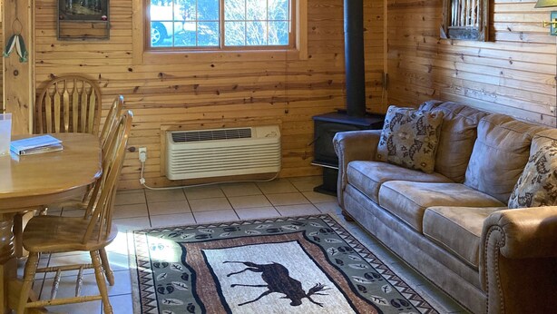 A cozy cabin interior with a wooden ceiling, a ceiling fan, a fireplace, a sofa, a dining table with chairs, and a window with a view of trees. - Wolf Pack place of Pomme de Terre Lake! (Pittsburg)