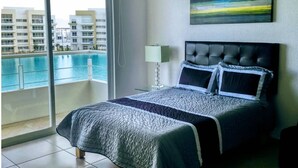 A bedroom with a bed, a nightstand, a lamp, and a view of a swimming pool and buildings. - Modern executive level suite in Metepec (Metepec)