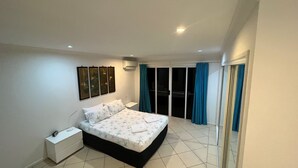 Un dormitorio con cama, mesita de noche, aire acondicionado instalado en la pared y puerta corrediza de vidrio que da a un balcón.