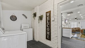 A reception area with a white brick wall, a welcome sign, a washing machine, and a view into a living room with a sofa and a coffee table.