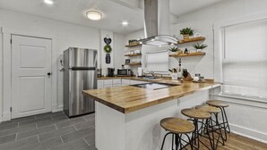 A modern kitchen with a central island, stainless steel appliances, wooden stools, and decorative shelves.