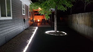 Een smal pad langs een boom en een houten hek, dat leidt naar een gebouw met een veranda en verlicht interieur.