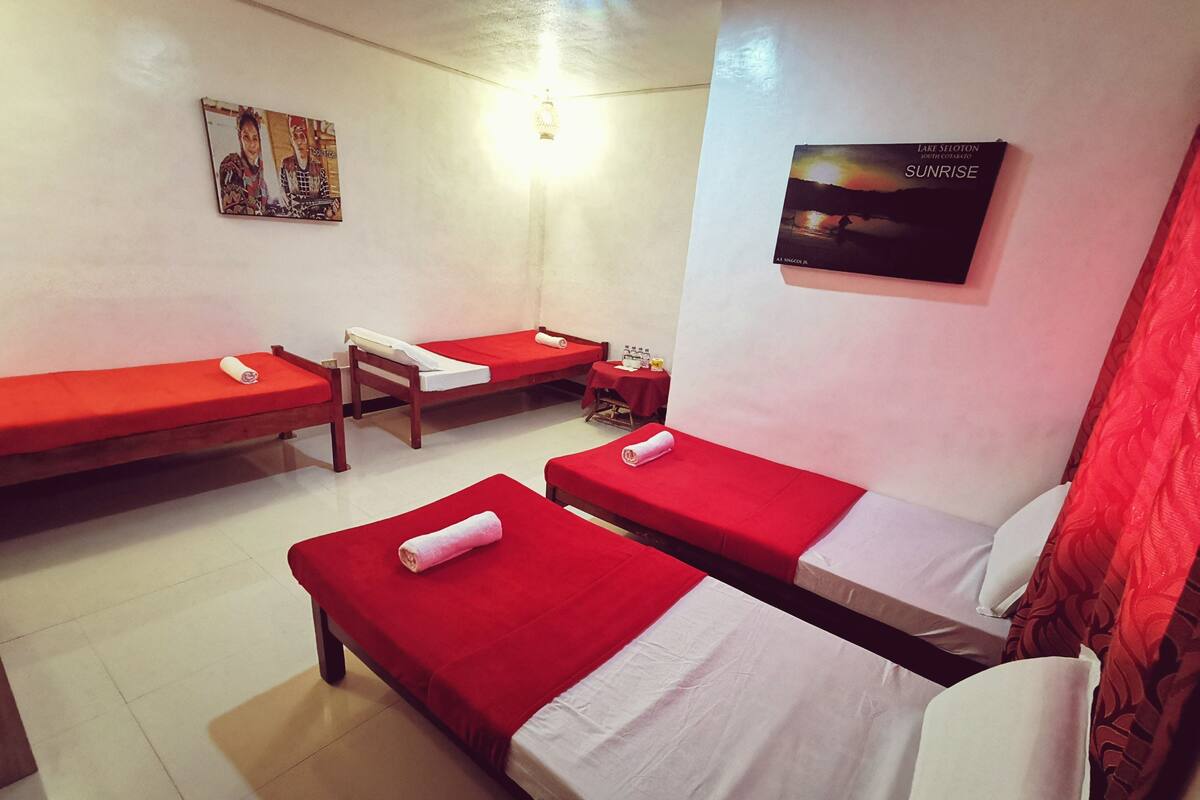 A room with four single beds, each with a red bedspread and white pillows. There is a framed picture on the wall and a poster of a sunset.