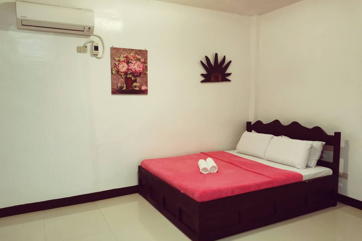 A single bed with a red bedspread and white pillows, a wall-mounted air conditioner, a floral painting, and a decorative wall piece.
