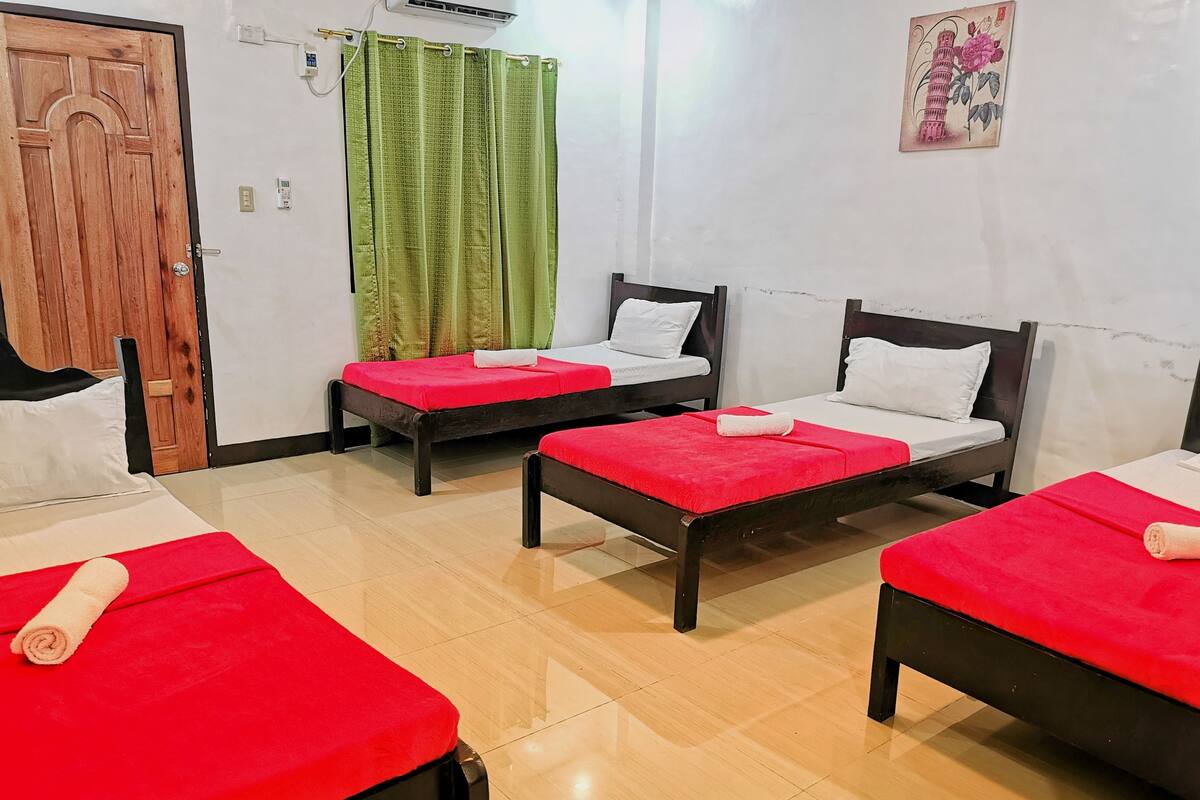 A room with three beds, each with a red bedspread and white pillows. There is a wooden door, a green curtain, and a wall-mounted air conditioner.