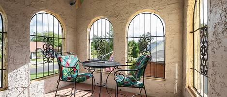Un patio soleado con tres ventanas de arco, una mesa redonda y dos sillas con tapicería colorida.