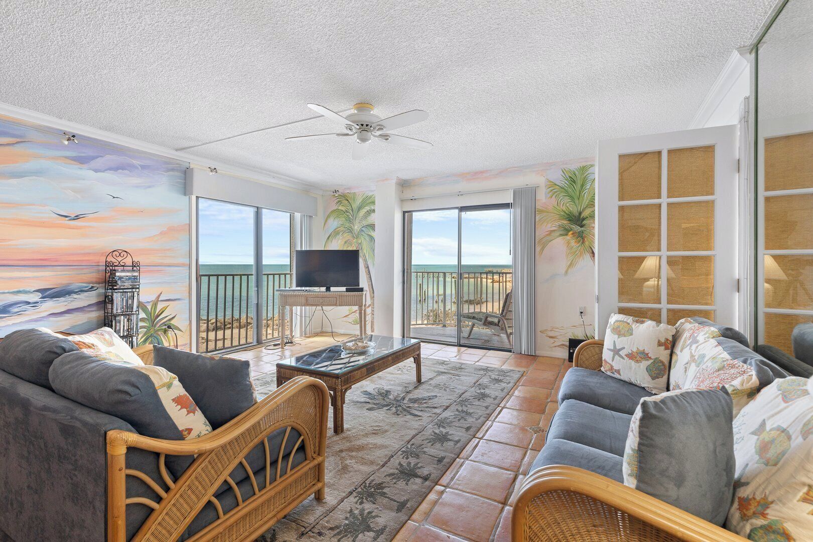 A spacious living room with a large TV, comfortable seating, and a balcony with a view of the ocean.